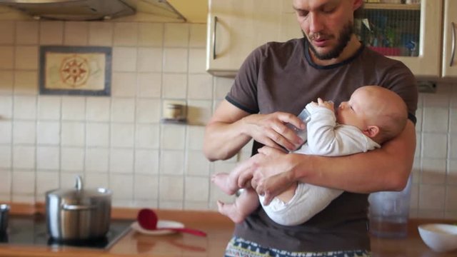 Stylish multitasking father feeds the baby in the kitchen and boils the soup, stirring it on the stove. A man has a stylish hairstyle and a modern beard