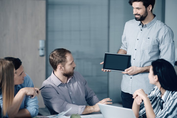 Share your experience. Cheerful young bearded man holding a tablet and presenting his project while working with his colleagues in one friendly team