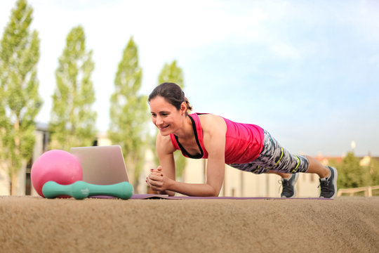 sporty and strong woman making planks infront of a laptop while watching online Fitness program on a mat in the park