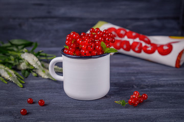 Ripe red currant berries in a white enamel cup and flowers on a dark background