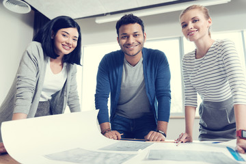 Look at us. International worker leaning on the table and keeping smile on his face while standing between near colleagues