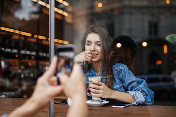 The boy makes a girl photo on the phone through the window in the cafe
