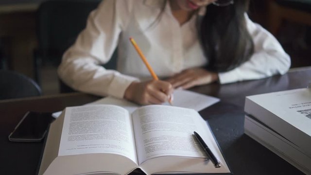 Book Is Lying On The Table And Female Student In White Shirt Making Conspect Of It In Her Notebook, Turning Pages By Her Hand With Manicure. Indoors.