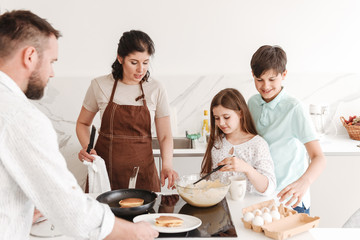 Lovely big family mother and father with two kids 8-10 cooking together, and frying pancakes on modern stove in kitchen at home