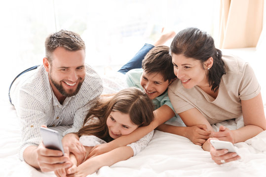 Portrait of joyful family with two children laughing, and taking selfie on smartphone while lying together on bed in apartment