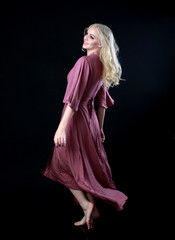 full length portrait of blonde girl wearing long purple dress. standing pose with back to the camera. black studio background.