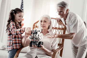 It is our work. Proud joyful grandfather and girl demonstrating robot pointing at it while  amazed grandma holding it in the rocking chair