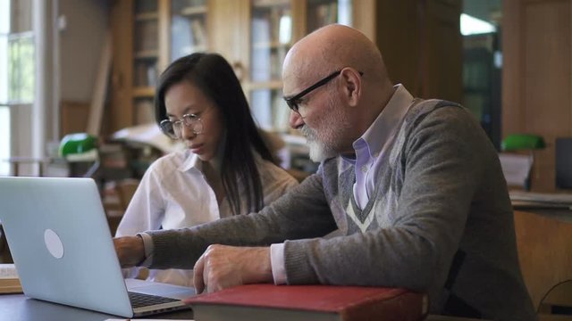 Mature professor and young female asian student working together on brand-new laptop in the library, discussing course paper. Indoors.