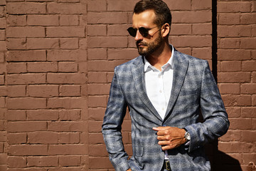 Portrait of sexy handsome fashion businessman model dressed in elegant checkered suit posing near brick wall on the street background. Metrosexual