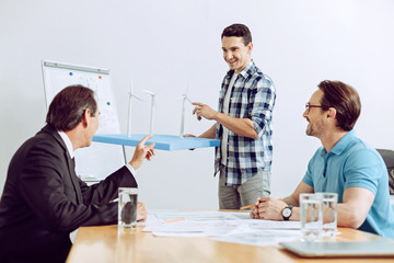 Exactly so. Smart experienced smiling engineer showing his wonderful project of a windmill generator to his clever attentive coworkers