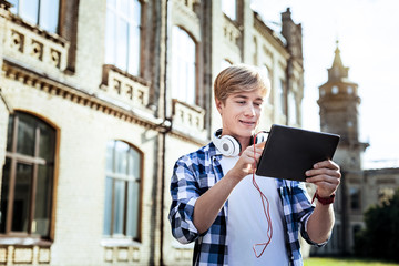 Let me check. Cheerful man having headphones around neck and looking at his gadget while standing near university