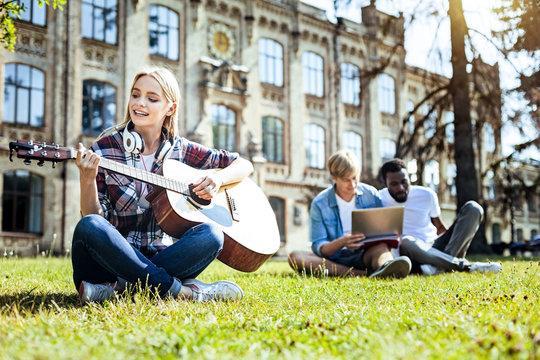 Favorite leisure activity. Attractive blonde lady in casual sitting on grass and smiling while playing guitar and relaxing outdoors.