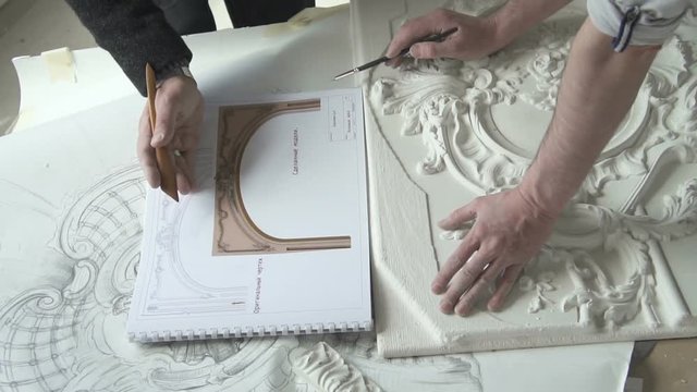 Male masters restorers working on restoration project at table in modern office, two architects are comparing plaster white decoration with colored drawings at desk in light workshop. Concept