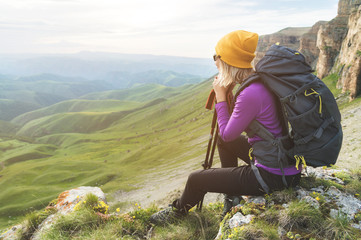 Smiling girl traveler in a yellow hat and a pair of sunglasses ssitting at the foot of epic rocks with a backpack next and looks away