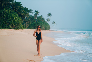 A stylish European strolls along the seashore, holds her hand on her thin waist, wears trendy sun glasses