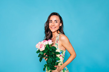 Happy and smiling young woman portrait with long curly hair in summer dress with bouquet of flowers in hands posing over blue background. Holidays and gifts concept