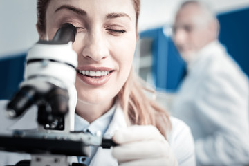 Professional biologist. Portrait of a positive delighted female scientist looking into the microscope and smiling while doing a research