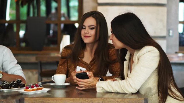 Two girls sitting in a cafe looking at the phone and discuss what they saw. A guy comes up and shows a funny picture on his teelphone