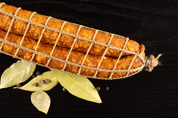 Group of two whole dry smoked ham sausage with bay laurel leaves and black pepper flatlay on black wood