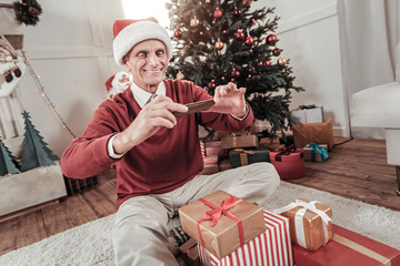 Obraz na płótnie Canvas Christmas mood. Happy pensioner keeping smile on his face and using telephone while sitting near presents