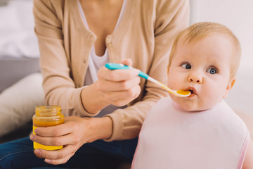 Delicious meal. Adorable little girl attentively looking into the distance while a kind calm mother carefully giving her a spoonful of baby food