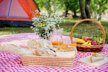 Picnic wicker basket with food, bread, fruit and orange juice on a red and white checked cloth in the field with green nature background. Picnic concept.