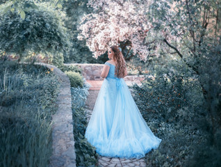 A big, beautiful, young woman in a luxurious blue dress with a long train is walking in a blooming...