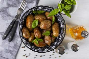 Baked potatoes in a black basket. Knife and fork. Olive oil, salt, pepper, basil. The background is white. Copy space. Top view. Horizontal shot.