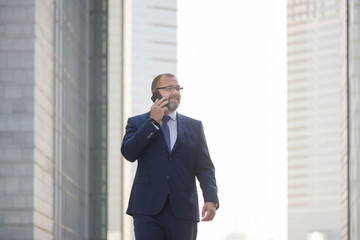 Closeup view of white middle age, bearded corporate businessman wearing a suit and tie with glasses, smiling  walking while talking on the phone
