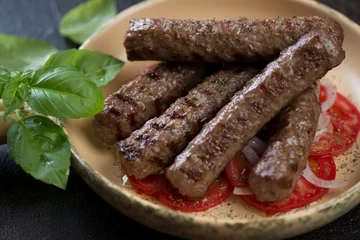 Fototapeten Closeup of grilled balkan cevapi or cevapcici sausages with tomatoes, onion and green basil leaves, selective focus © Nickola_Che