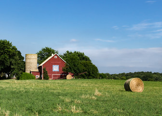 Bright red barn with barn quilt and silo against a bright blue summer sky. Large round hay bales are in the field in front of the barn. Concepts of family farm, farming, agriculture, harvest, summer © Kimberly Boyles