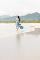 Fashion outdoor photo of gorgeous young lady in long blue dress walking barefoot in water at sea shore. Summer sunny day and holiday concept, seascape with girl, beach, beautiful waves, blue water.