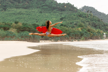 Outdoor photo of gorgeous young woman in fashion bikini doing jump split leap with red surfboard. Summer sunny day sport surfing concept, seascape with girl, beach, beautiful waves, blue water. - 214121949