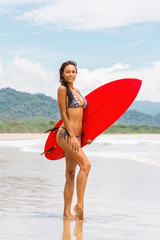 Naklejka na ściany i meble Lifestyle photo of young lady in fashion bikini with red surfboard smile to camera. Amazing beach surf style girl, windy hair, toned, sugaring legs, beach wear, tender skin. Good summer day concept.