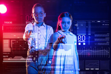 Touching the screen. Calm modern kids standing in a futuristic classroom and thoughtfully pointing to a transparent screen