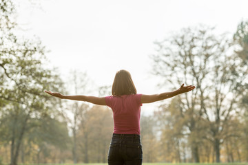 View from behind of a young woman standing in park with her arms spread widely