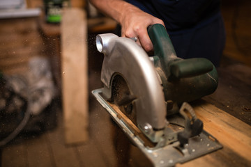 A young male carpenter-carpenter saws a modern circular saw with a wooden board in the workshop, wooden sawdust flying in the sides. to plan a tree.