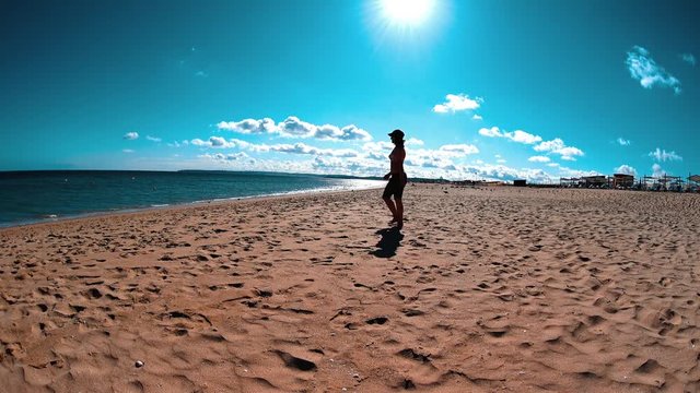 Girl in swimsuit, denim shorts and snapback walking across the beach to water line, summer sunny day at vacation
