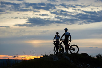 Fototapeta na wymiar Back view of two young cyclists with mountain bikes standing on top of a cliff with beautiful scenery of hills and sky at sunset. The concept of a healthy lifestyle