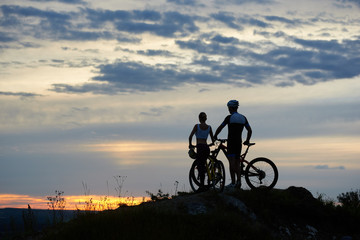 Obraz premium Back view of two young cyclists with mountain bikes standing on top of a cliff with beautiful scenery of hills and sky at sunset. The concept of a healthy lifestyle