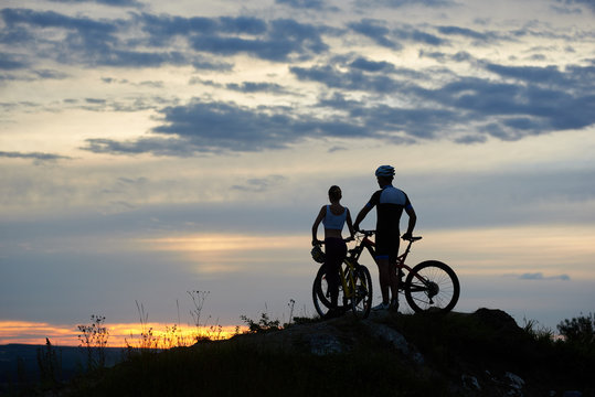 Back view of two young cyclists with mountain bikes standing on top of a cliff with beautiful scenery of hills and sky at sunset. The concept of a healthy lifestyle