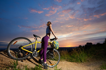 Fototapeta na wymiar Slender woman wearing violet leggings, posing with her back and buttocks turned to camera. Athletic cyclist standing with yellow bicycle, looking at sunset and beautiful landscape.