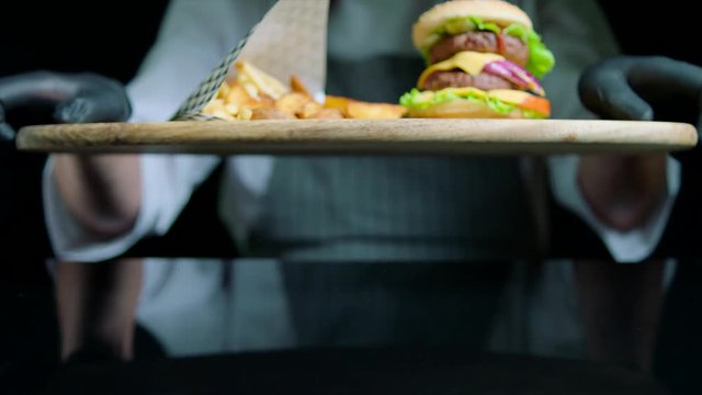 Chef is serving set of tasty burger, french fries, beer on the wooden tray on the black background