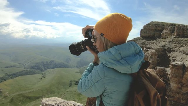 Blonde girl photographer in the cap takes a photo on her digital camera with a background of rocks in the Caucasus