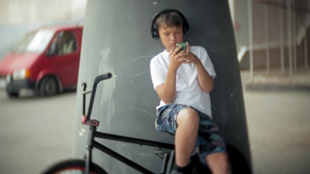 The boy sits on a BMX bike and listens to music from a smartphone