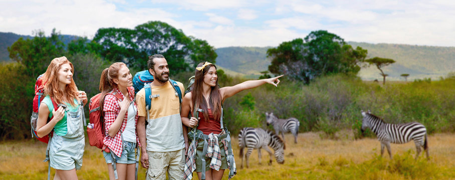 travel, tourism, hike and adventure concept - group of smiling friends with backpacks pointing finger to something over zebras in african savannah background