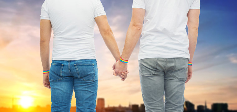 lgbt, same-sex relationships and homosexual concept - close up of male couple wearing gay pride rainbow awareness wristbands holding hands over sunset in tallinn city background