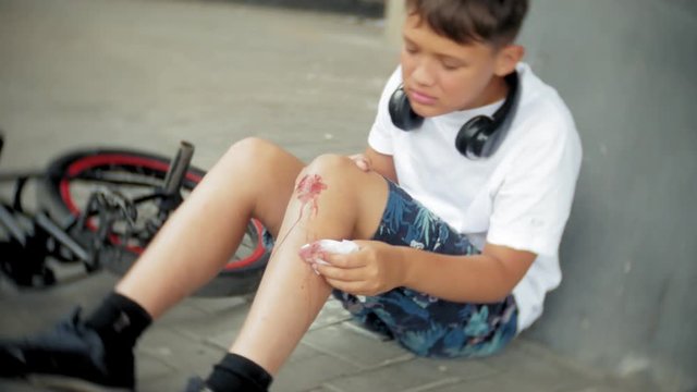 A young boy sits in a park after falling from a bike, calms the pain in his knee, is a dangerous bike ride