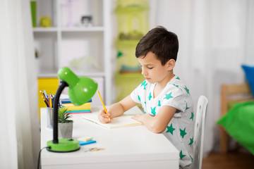 education, childhood and school concept - little boy sitting at desk and writing to notebook at home