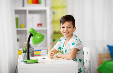 education, childhood and school concept - happy little boy sitting at desk and writing to notebook...